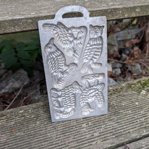 Puede incluir: Un molde de metal rectangular vintage con asa, con diseños de aves en relieve. El molde incluye un búho, un pato y otras figuras de aves. El molde de color plateado descansa sobre una superficie de madera desgastada.