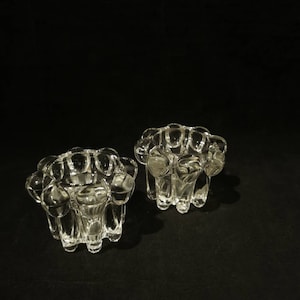 Könnte beinhalten: Zwei durchsichtige Glas-Kerzenhalter mit einem einzigartigen, strukturierten Design. Jeder Halter hat eine Reihe von abgerundeten, blasenartigen Formen um den Rand und die Basis, wodurch ein dekoratives und elegantes Aussehen entsteht. Die Kerzenhalter sind vor einem dunklen Hintergrund platziert.
