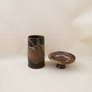 Könnte beinhalten: Eine Keramikvase und eine kleine Schale mit Sockel. Die Vase ist zylindrisch, mit einem dunkelbraunen Innenraum und einer schwarzen Außenseite mit braunen und weißen abstrakten Mustern. Die Schale ist braun mit weißem Akzent.