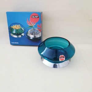 Op de afbeelding: Een turquoise glazen kom met een zilveren voet. De kom staat op een wit oppervlak. De kom heeft een label met de tekst "Quist Presena".