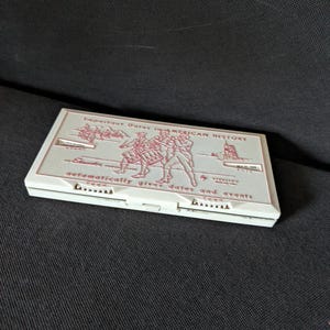 Peut inclure: Un étui rectangulaire blanc avec du texte et des illustrations en relief rouge. Le texte indique "Important Dates in American History" et "automatically gives dates and events". L'étui présente des images de soldats et d'un bâtiment.
