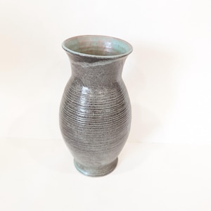 Pode incluir: Um vaso de cerâmica cinza com um corpo texturizado e canelado e uma borda alargada. A borda interna tem um esmalte turquesa. O vaso tem uma base redonda e é adequado para exibir flores ou como peça decorativa.