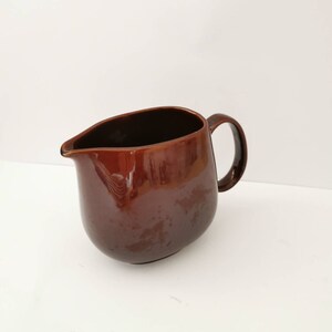 May include: A brown ceramic pitcher with a rounded body and a handle. The pitcher is made of a smooth, glossy ceramic.