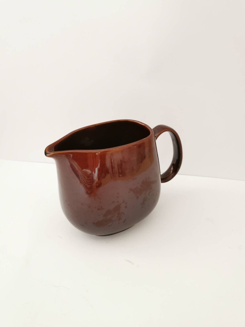 May include: A brown ceramic pitcher with a rounded body and a handle. The pitcher is made of a smooth, glossy ceramic.