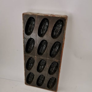 Könnte beinhalten: Eine antike Holzform mit 12 einzelnen Vertiefungen zum Backen von kleinen Kuchen oder Gebäck. Die Form ist dunkelbraun mit einer abgenutzten Oberfläche und hat eine rechteckige Form.