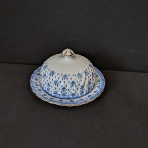 以下が含まれることがあります： 青い花柄と渦巻き状のドーム型蓋が付いた白い磁器製のバターケース。皿の縁はスカラップ状で金色の縁取りが施されており、蓋の頂上には小さな金色のつまみが付いています。デザインには繊細な青い植物のモチーフが描かれています。