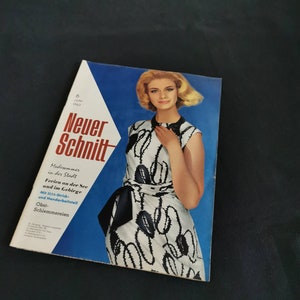 Pode incluir: Uma capa de revista vintage com uma mulher em um vestido estampado em preto e branco. O título da revista "Neuer Schnitt" é exibido em vermelho e branco, com texto adicional em alemão. O fundo é azul e a revista está em ângulo.