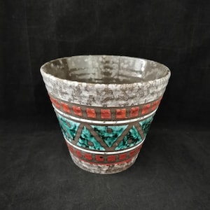 Op de afbeelding: Een keramische plantenbak met een getextureerde, gespikkelde grijze rand en een patroonlichaam. De body heeft banden van rode vierkanten en turquoise driehoeken, gescheiden door bruine lijnen. De plantenbak heeft een brede opening en een donker interieur.