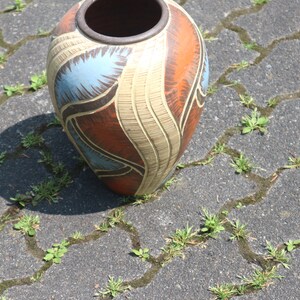 May include: A large, ceramic vase with a brown, orange, blue, and white abstract design. The vase is sitting on a gray, textured surface.