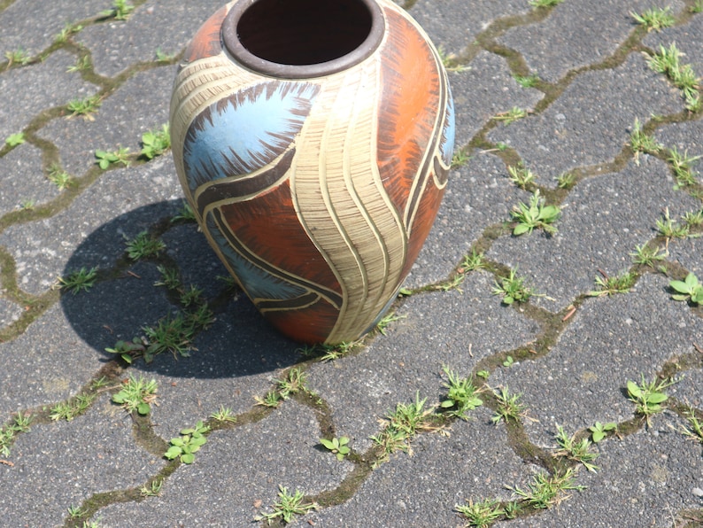 May include: A large, ceramic vase with a brown, orange, blue, and white abstract design. The vase is sitting on a gray, textured surface.