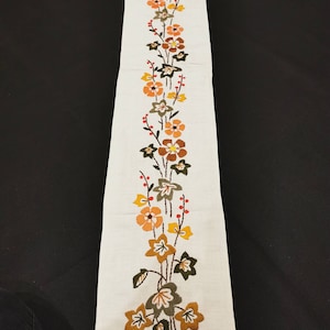 Könnte beinhalten: Cremefarbener, bestickter Tischläufer mit vertikalem Blumenmuster. Die Blumen sind in Orange-, Braun- und Gelbtönen gehalten, mit grünen Blättern und roten Beerenakzenten. Der Läufer ist rechteckig und scheint aus Leinen oder einem ähnlichen Stoff zu sein.