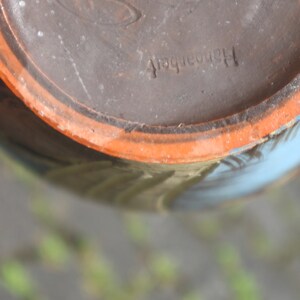 May include: Close-up of the bottom of a brown ceramic pot with a light orange rim. The pot has a blue and white design and the text "Handarbeit" is stamped on the bottom.