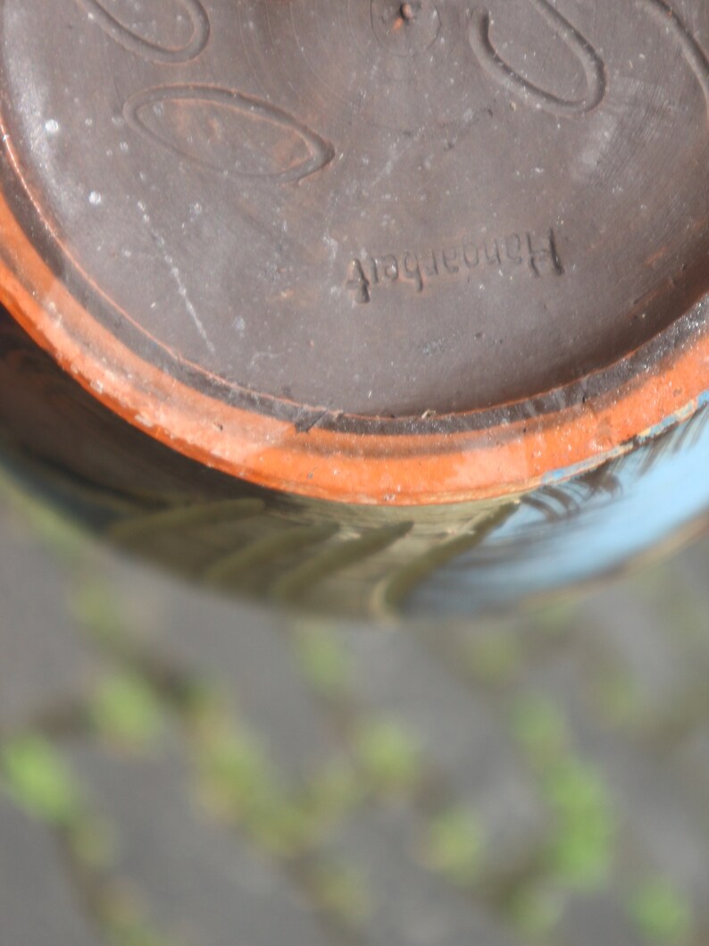 May include: Close-up of the bottom of a brown ceramic pot with a light orange rim. The pot has a blue and white design and the text "Handarbeit" is stamped on the bottom.
