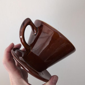 Op de afbeelding: Een bruine keramische kop en schotel set. De kop heeft een gebogen oor en een glanzende afwerking. De ronde schotel past bij de kleur van de kop. De set wordt schuin gehouden tegen een effen witte achtergrond.