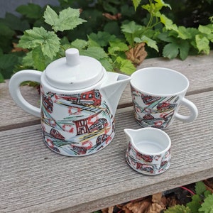 May include: A white tea set with a red, green and blue pattern of trains. The set includes a teapot, a cup and a creamer.
