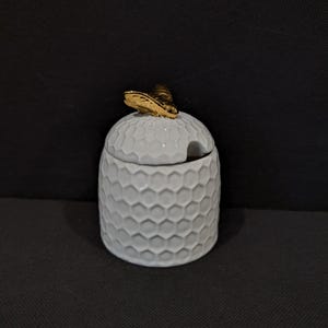 Könnte beinhalten: Ein weißer Honigtopf aus Keramik mit Wabenmuster und einem goldfarbenen, bienenförmigen Deckelgriff. Der Topf hat eine kleine Öffnung für einen Löffel. Das Design ist schlicht und elegant.