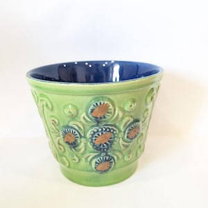 Op de afbeelding: Een groene keramische bloempot met een blauwe binnenkant. De buitenkant heeft reliëf bloemmotieven met bruine en blauwe accenten. De pot heeft een licht uitlopende vorm en is geschikt voor kamerplanten.
