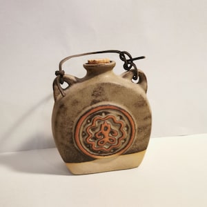 Könnte beinhalten: Eine braune und beigefarbene Keramikflasche mit einem Korkverschluss und einem Lederriemen als Henkel. Die Flasche hat ein kreisförmiges Design mit einem braunen und orangefarbenen Muster.