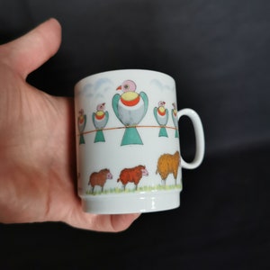 Könnte beinhalten: Weiße Keramiktasse mit einem bunten Vogeldesign und einer Reihe von Schafen. Die Vögel sind grün, gelb, rosa und orange. Die Schafe sind braun, weiß und orange.