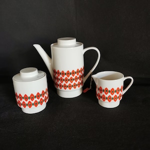 Könnte beinhalten: Ein weißes Keramik-Tee-Set mit einem geometrischen Muster in Rot, Grün und Weiß. Das Set enthält eine Teekanne, eine Milchkännchen und eine Zuckerdose.