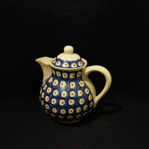 Op de afbeelding: Een keramische theepot met een blauw en wit bloemmotief. De theepot heeft een gebogen tuit, een handvat en een deksel met een kleine knop. Het ontwerp heeft blauwe stippen met gele centra op een witte achtergrond.