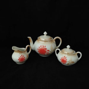 Op de afbeelding: Een wit porseleinen theeservies met een bloemenmotief. De set bevat een theepot, een roomkan en een suikerpot. De bloemen zijn rood en bruin.