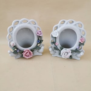 Könnte beinhalten: Zwei weiße Keramik-Bilderrahmen mit Blumenmuster. Jeder Rahmen hat eine weiße ovale Öffnung und ist mit rosa, weißen und blauen Blumen verziert.