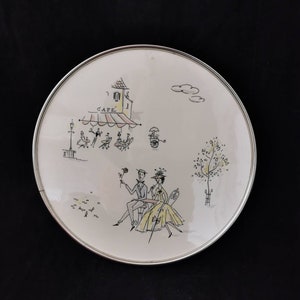 Puede incluir: Un plato decorativo redondo con borde plateado. El plato presenta una ilustración caprichosa de una escena de café con gente, un letrero de café y una pareja sentada a una mesa. La ilustración es en negro, amarillo y verde.