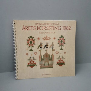 Puede incluir: Un libro titulado "Årets Korssting 1982" con el subtítulo "Nye Navneklude" de Ida Winckler. La portada presenta un diseño de punto de cruz de una casa con pájaros, flores y corazones.