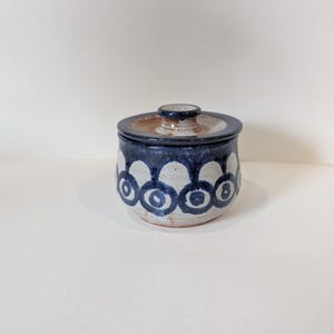 Peut inclure: Un pot en céramique avec un couvercle. Le pot est cylindrique avec une bande bleu foncé en haut et un motif répétitif de cercles bleus et blancs sur le corps. Le couvercle est de couleur plus claire avec un bord marron.