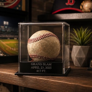 May include: A baseball in a clear display case. The baseball has red stitching and is displayed on a black base with the text "GRAND SLAM APRIL 23, 2025 417 FT." A baseball glove, cap, and trophy are in the background.