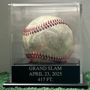 May include: A baseball in a clear display case. The baseball is off-white with red stitching. A black plaque below the ball reads "GRAND SLAM APRIL 23, 2025 417 FT."