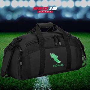 May include: Black duffel bag with a green logo and the name "MICHAEL" in green. The bag has a shoulder strap and top handles. The background is a green field with stadium lights.