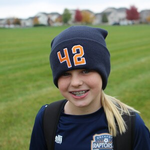 May include: A young person wearing a blue beanie with the number 42 on it. They are wearing a blue shirt with the words "Rockford Raptors" on it. They are smiling and have braces.