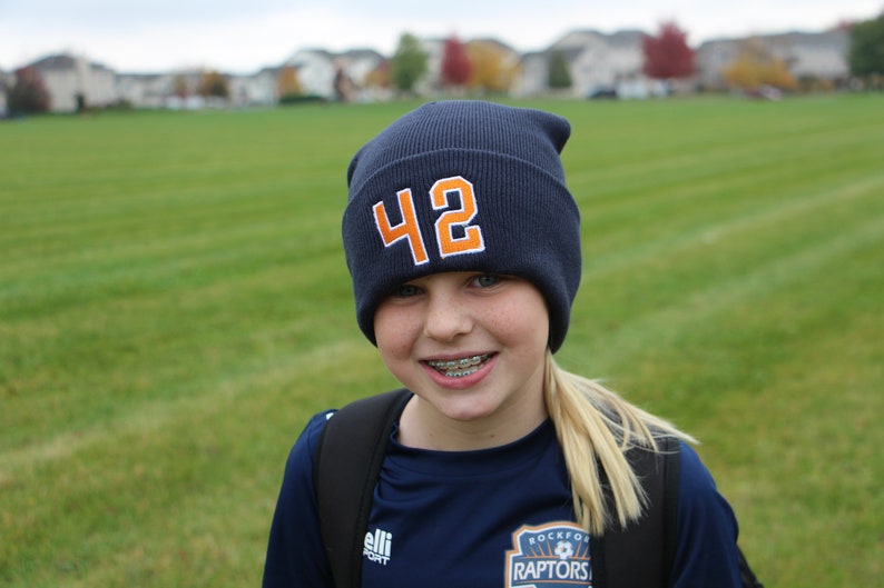May include: A young person wearing a blue beanie with the number 42 on it. They are wearing a blue shirt with the words "Rockford Raptors" on it. They are smiling and have braces.