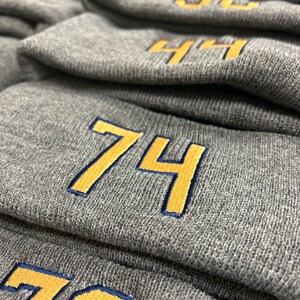 May include: Stack of gray knit beanies with yellow numbers embroidered on the front. The numbers are 74 and 76.