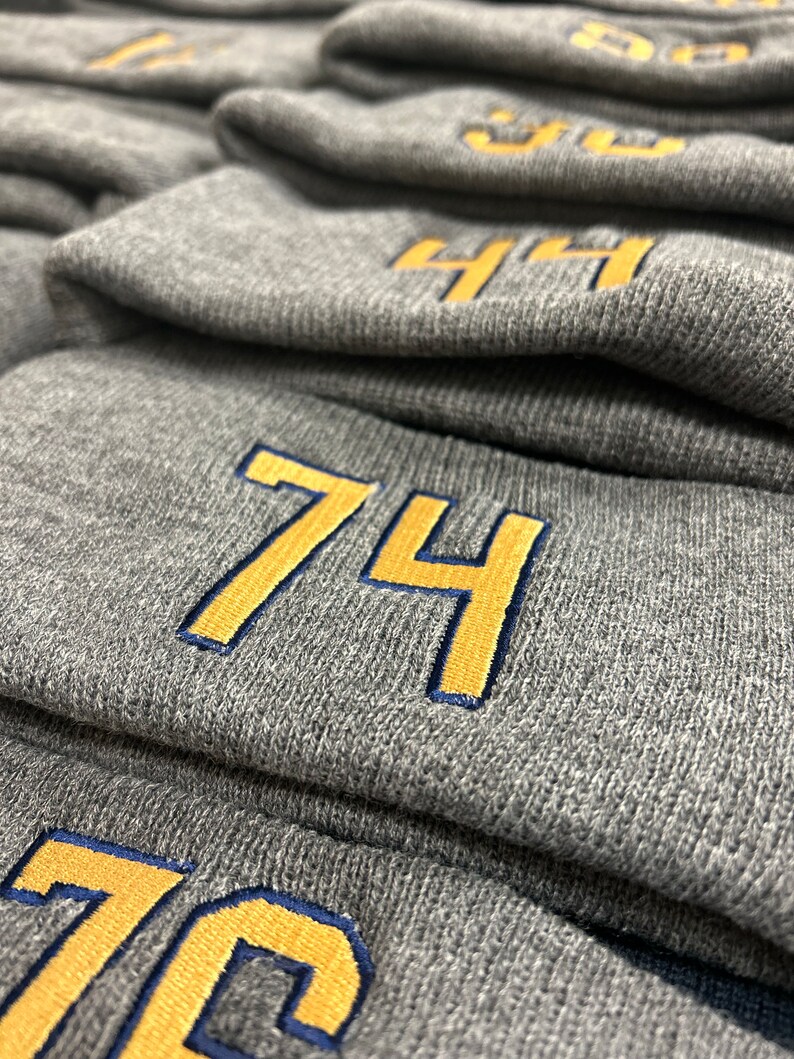 May include: Stack of gray knit beanies with yellow numbers embroidered on the front. The numbers are 74 and 76.