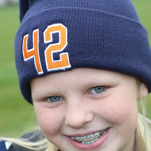May include: A young person wearing a blue beanie with the number 42 embroidered on it in orange. They are smiling and have braces on their teeth.