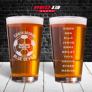 May include: Two pint glasses filled with beer, featuring custom text. One glass has a soccer ball graphic with "Coach George 2025 Blue Devils." The other glass lists names. The glasses are on a white wooden surface.