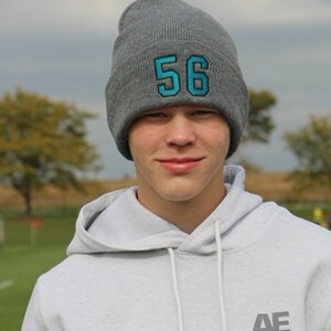 May include: A young person wearing a gray hooded sweatshirt with the text "AE 1977" on the front. They are also wearing a gray beanie with the number "56" in blue on the front.
