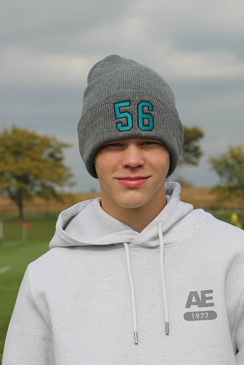 May include: A young person wearing a gray hooded sweatshirt with the text "AE 1977" on the front. They are also wearing a gray beanie with the number "56" in blue on the front.