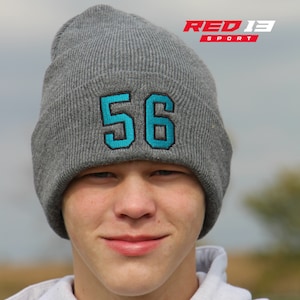 May include: A gray knit beanie with a turquoise number 56 embroidered on the front. The beanie is worn by a person with a light complexion and short brown hair. The beanie is pulled down over the person's forehead.