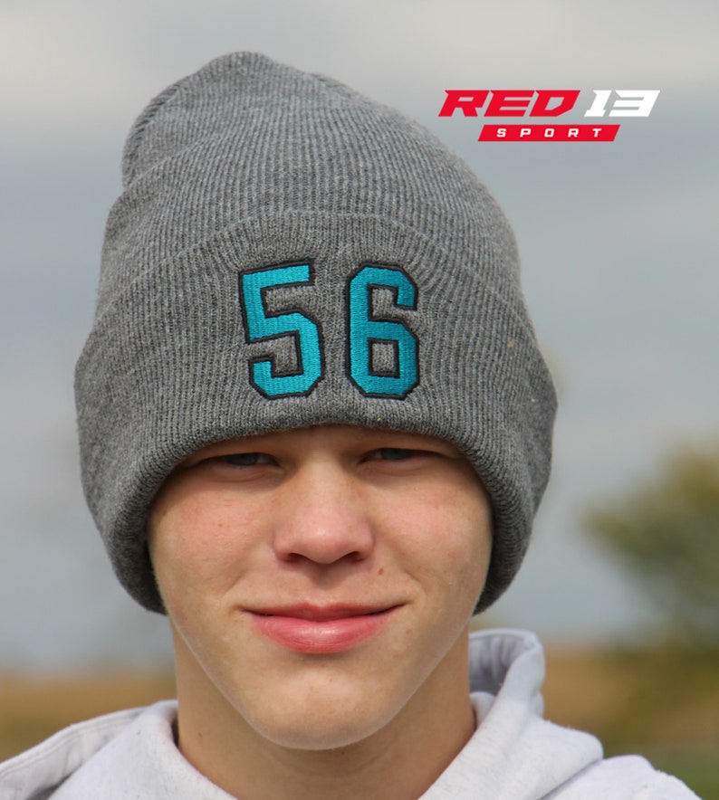 May include: A gray knit beanie with a turquoise number 56 embroidered on the front. The beanie is worn by a person with a light complexion and short brown hair. The beanie is pulled down over the person's forehead.
