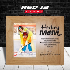Op de afbeelding: Een houten fotolijst met een foto van een moeder en kind op een ijshockeybaan. De lijst heeft een gegraveerde inscriptie die "Hockey Mom" met een hockeypuck en stick toont. De inscriptie gaat verder met "Through all the cold practices, home and away games, and long weekends at tournaments, you were always there. Thank you! Love, Bryant & Connor."