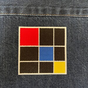 May include: A square patch with a white border and a grid of nine squares. The top left square is red, the middle square is blue, and the bottom right square is yellow. The remaining squares are black.