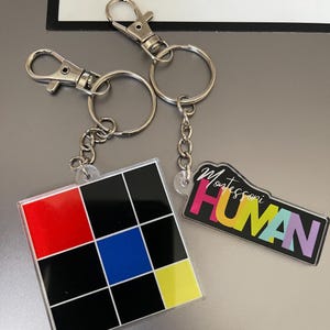 Pode incluir: Dois chaveiros com pingentes de acrílico coloridos. Um pingente é um quadrado com um design de blocos de cores vermelho, preto, azul e amarelo. O outro pingente é um retângulo preto com o texto "Montessori HUMAN" em cores do arco-íris.