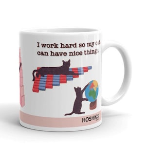 Puede incluir: Taza de cerámica blanca con una ilustración de un gato negro y el texto "Trabajo duro para que mi gato tenga cosas bonitas". El gato está tumbado en una gráfica de barras con barras rojas y azules. Un segundo gato negro está de pie en una pequeña plataforma sosteniendo un globo terráqueo. La taza tiene una banda rosa alrededor de la parte inferior y el texto "Hoshiko" en negro.