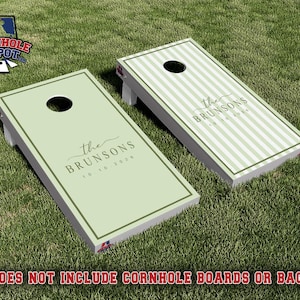 Könnte beinhalten: Zwei rechteckige Cornhole-Boards mit hellsalbeigrüner Oberfläche. Ein Board ist einfarbig salbeigrün mit dunkelgrünem Rand, das andere hat vertikale salbeigrüne Streifen. Beide tragen den Schriftzug the BRUNSONS 10 10 2026.