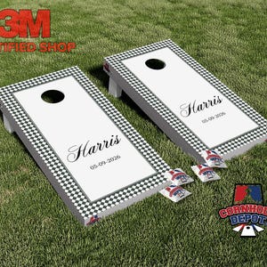 May include: Two cornhole boards with a white surface and a black and white checkered border. Each board has a centered hole and the name "Harris" with the date "05-09-2026". Several bean bags are visible.