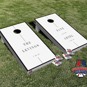 Könnte beinhalten: Zwei weiße Holz-Cornhole-Bretter mit schwarzem Rahmen und schwarzen Linien in der Mitte. Die Bretter sind mit dem Text "The Levings 7.28.23" und "Zane and Ariel 7.28.23" personalisiert.